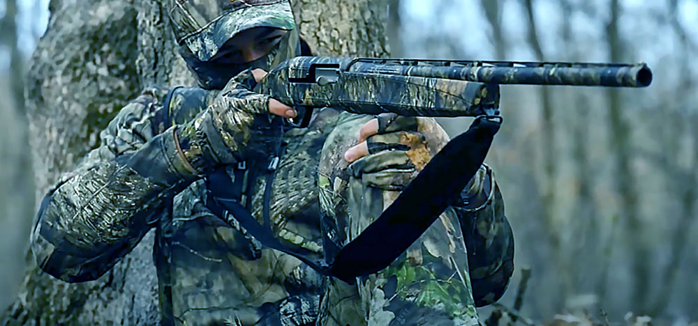 A man dressed in camouflage gear stands confidently, holding a shotgun in a poised position, ready for action